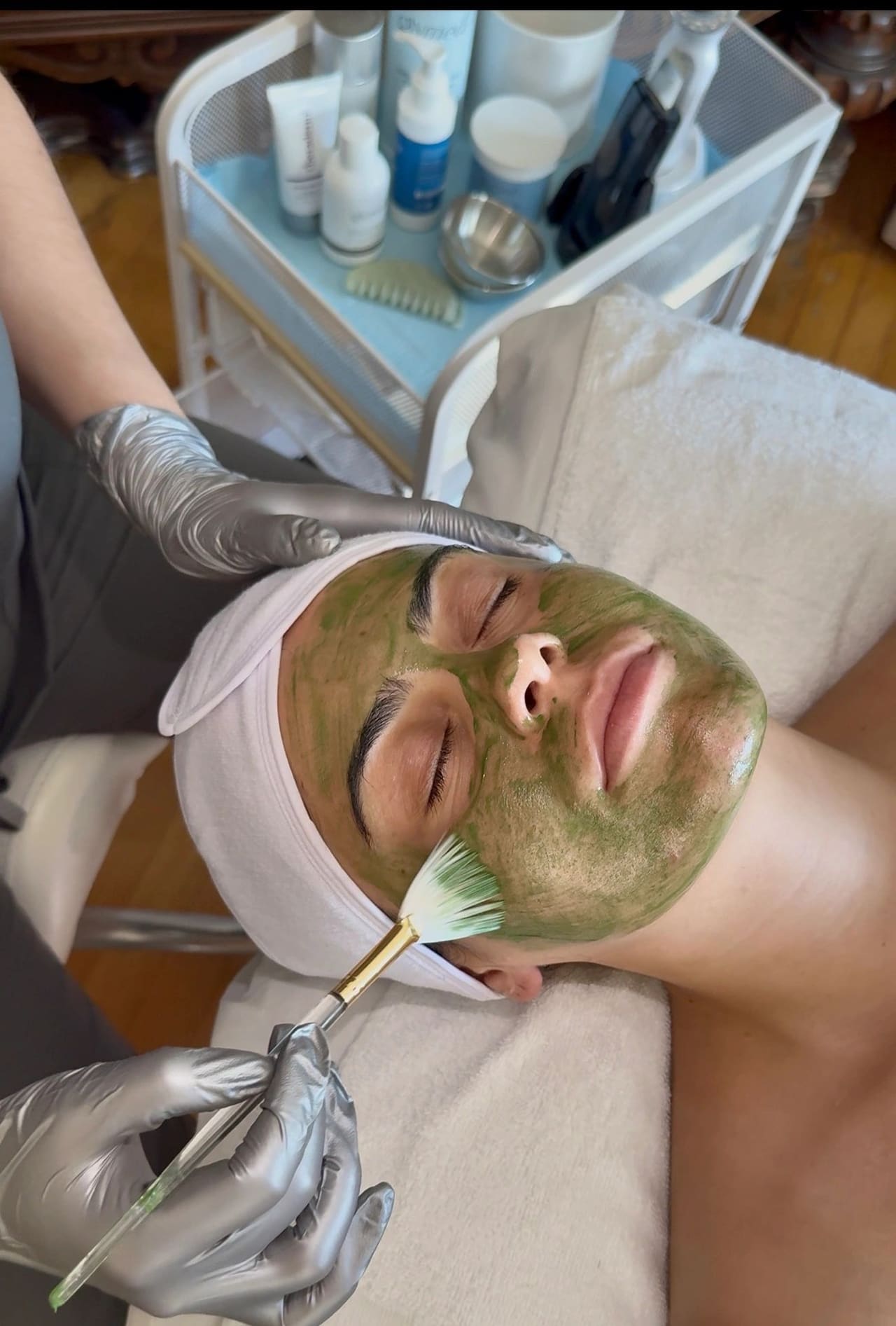 A green treatment mask being applied with a fan brush during a facial, product trolley alongside